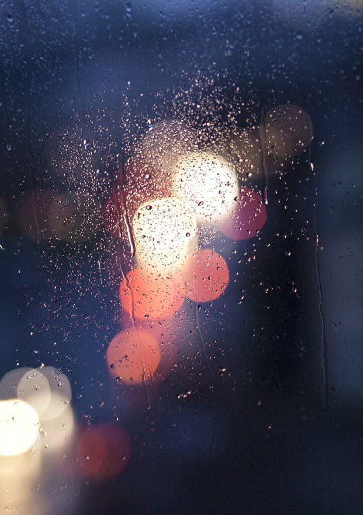A close-up of a rain-covered window at night. Raindrops cling to the glass, with blurred, glowing lights of cars in the background, creating a soft bokeh effect in warm hues of red, orange, and white.